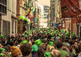 Crowded Dublin street during St Patrick’s Day with people wearing green hats and clothing, viewed from behind, walking between pubs and buildings decorated with Irish flags and festive banners.