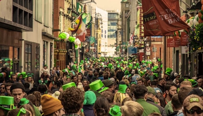 Crowded Dublin street during St Patrick’s Day with people wearing green hats and clothing, viewed from behind, walking between pubs and buildings decorated with Irish flags and festive banners.