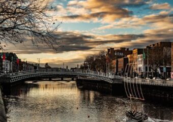 Best Free Things to Do in Dublin. A vibrant sunset view of the River Liffey in Dublin, featuring the iconic Ha'penny Bridge in the foreground with warm orange skies, autumn trees and traditional Georgian buildings lining the waterfront.