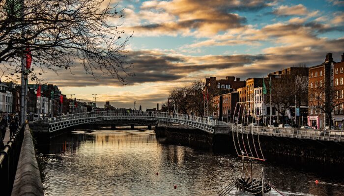 Best Free Things to Do in Dublin. A vibrant sunset view of the River Liffey in Dublin, featuring the iconic Ha'penny Bridge in the foreground with warm orange skies, autumn trees and traditional Georgian buildings lining the waterfront.